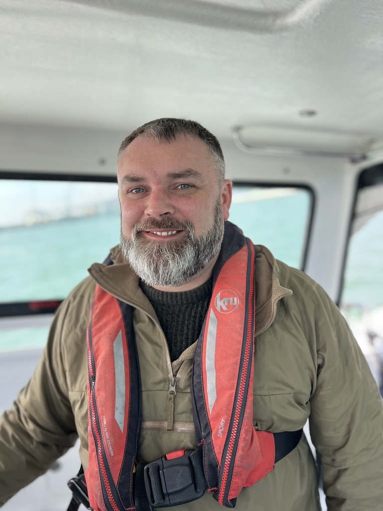 Gentleman with a beard smiling wearing a green jacket and a red lifejacket