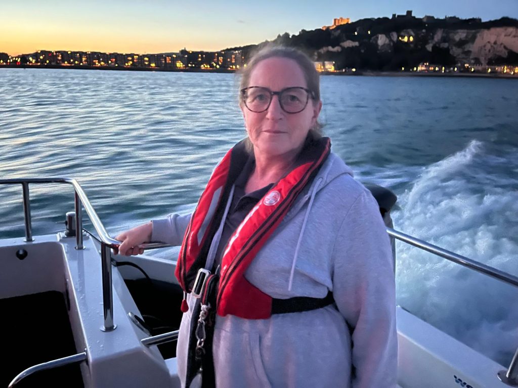 Lady with long hair in a pony tail, wearing glasses and a red life jacket. She is standing on a powerboat with a sunset behind her.